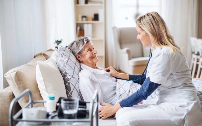 A young health visitor examining a sick senior woman lying in bed at home with stethoscope.