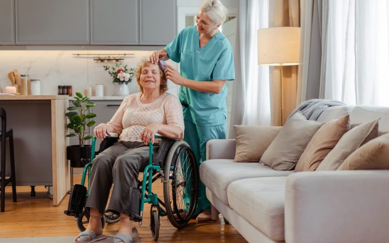 Nurse in scrubs caring for a happy senior woman in a wheelchair at home, brushing her hair in a cozy living room
